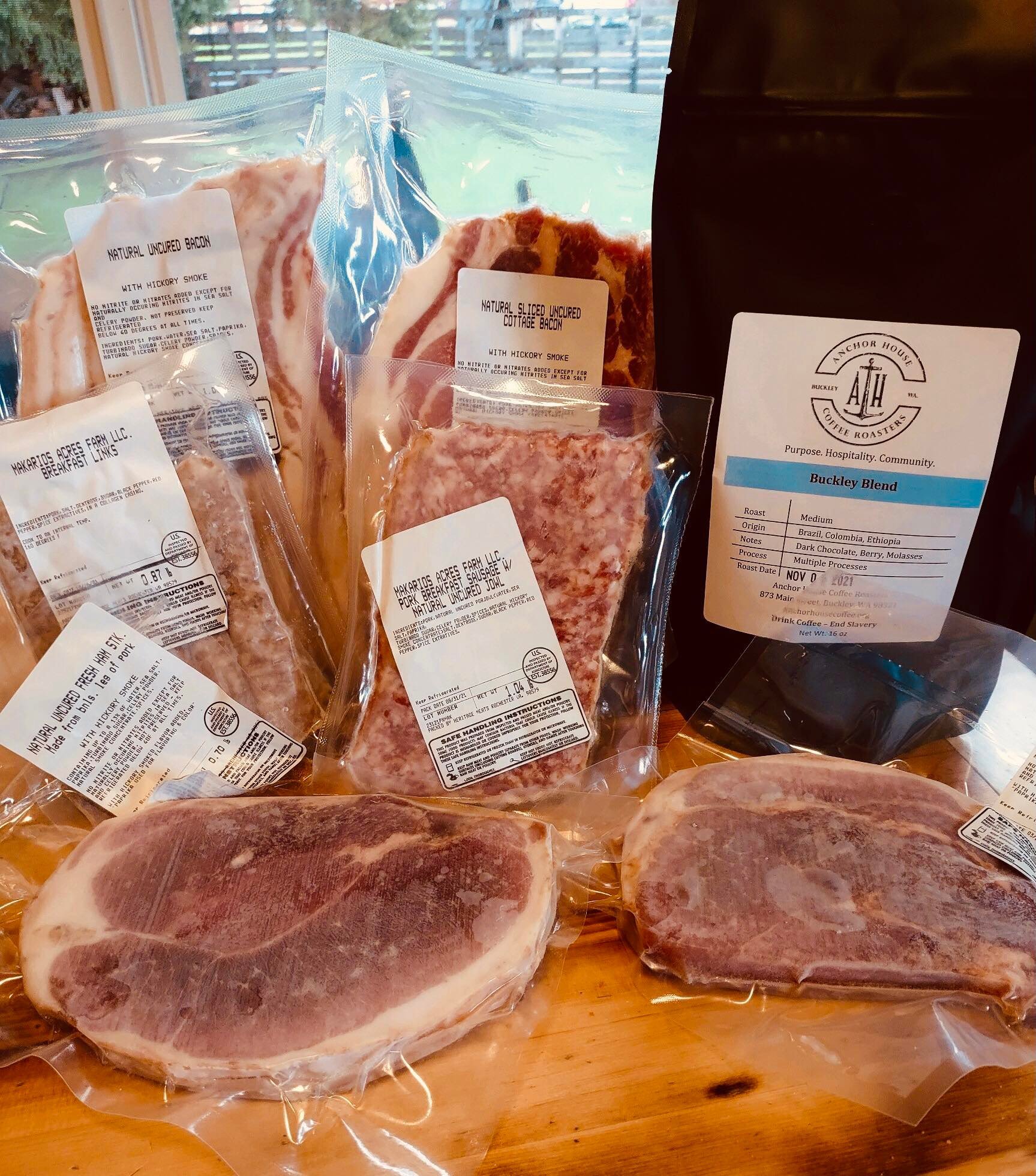 Assorted packages of natural, uncured bacon and pork cuts from Wakarius Acres Farm, featuring hickory-smoked cottage bacon and a variety of labeled meats, alongside a bag of Buckley Blend coffee from Anchor House Coffee Roasters, highlighting its origin and tasting notes.