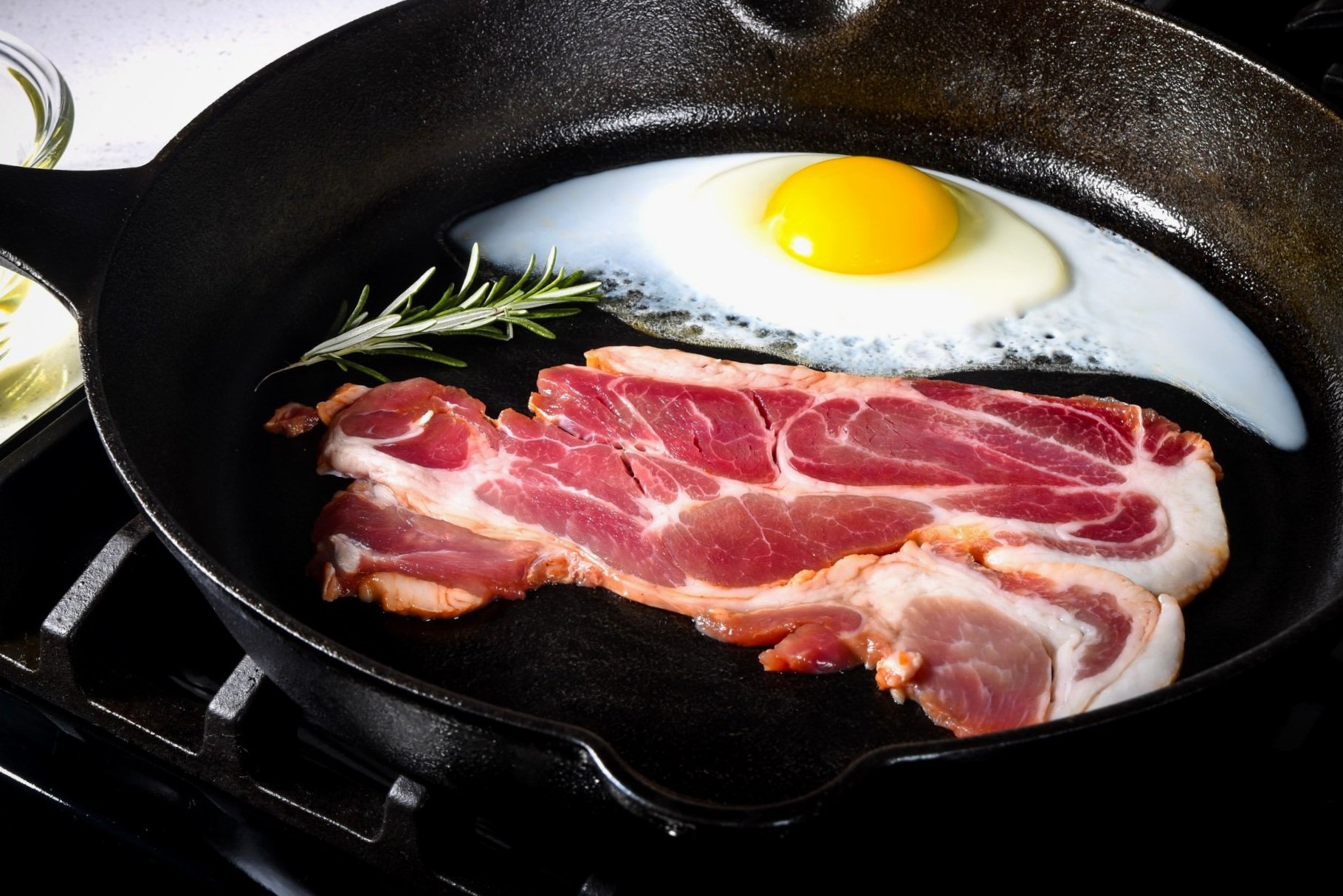 Cooked bacon and a sunny-side-up egg in a cast iron skillet, garnished with a sprig of rosemary, showcasing a delicious breakfast preparation.
