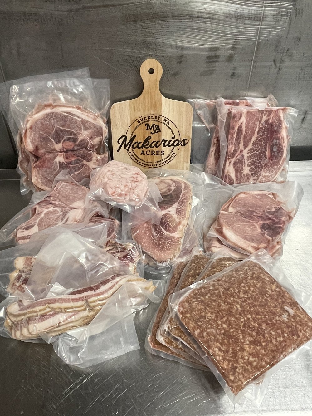 Assorted vacuum-sealed cuts of meat including steaks, bacon, and ground meat from Makario's Acres, displayed with a wooden cutting board featuring the brand's logo. Ideal for meat lovers seeking high-quality, locally sourced products.