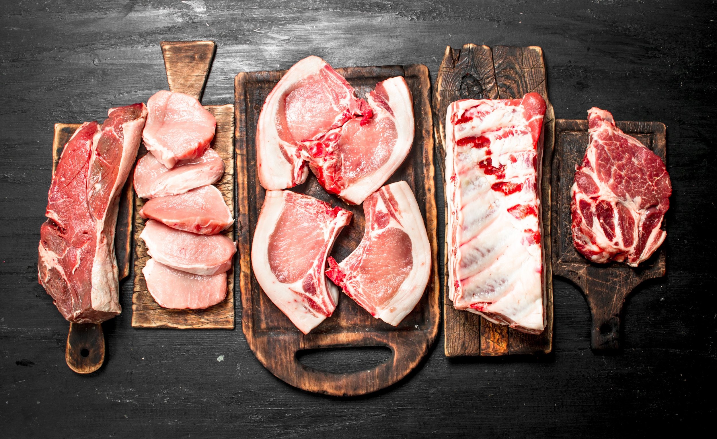 Assorted raw meats displayed on wooden cutting boards, including pork chops, pork loin, beef ribs, and steak cuts, set against a dark background. Ideal for meat preparation and culinary inspiration.