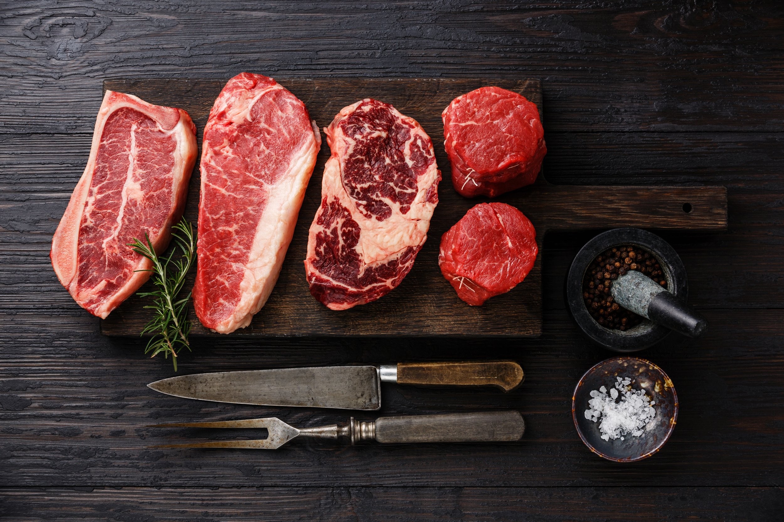 Assorted cuts of raw beef, including ribeye, sirloin, and tenderloin, arranged on a wooden cutting board with a sprig of rosemary, alongside kitchen utensils and spices.