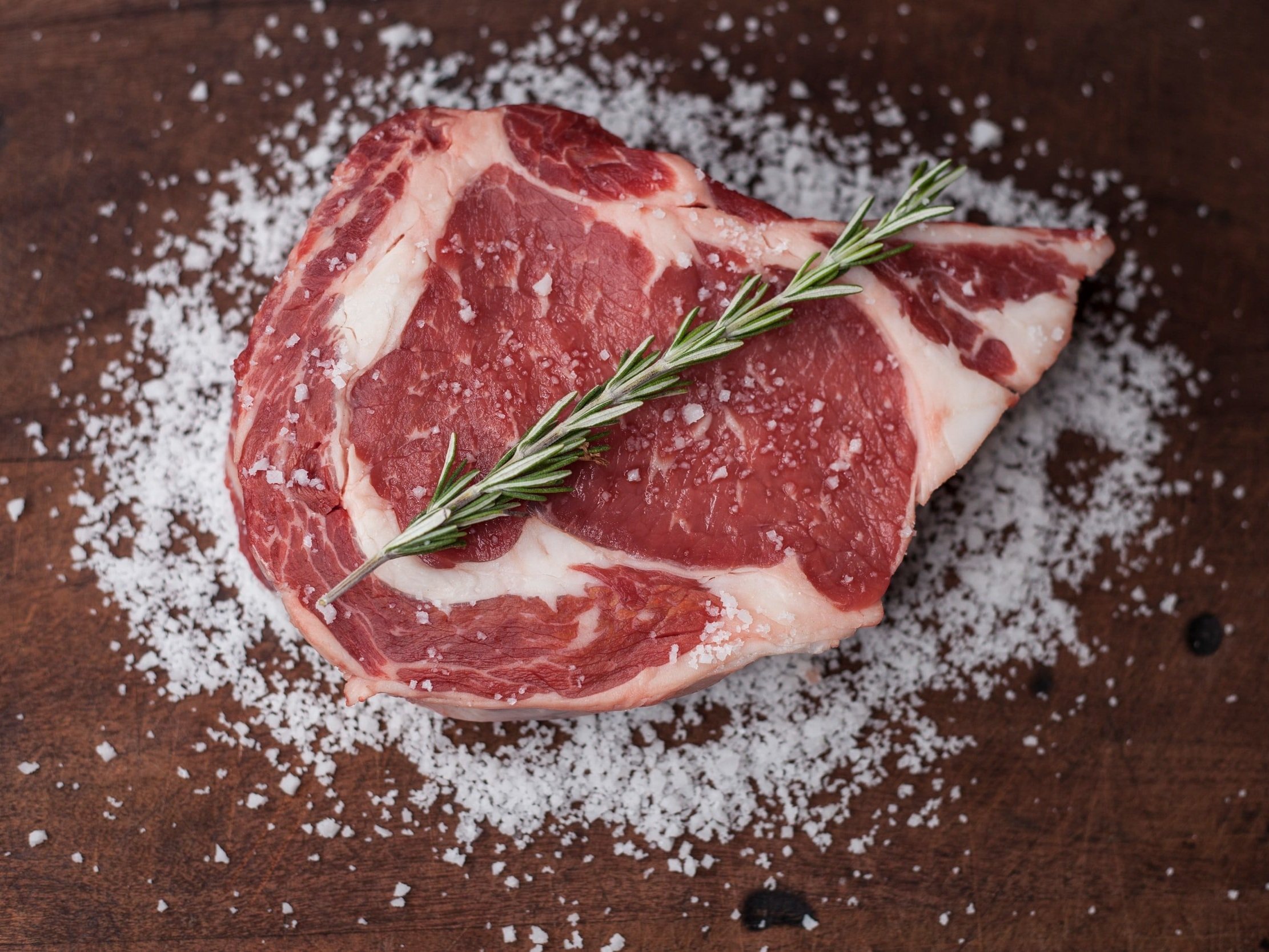 Fresh ribeye steak garnished with a sprig of rosemary, surrounded by coarse sea salt on a wooden cutting board. Ideal for grilling or roasting.